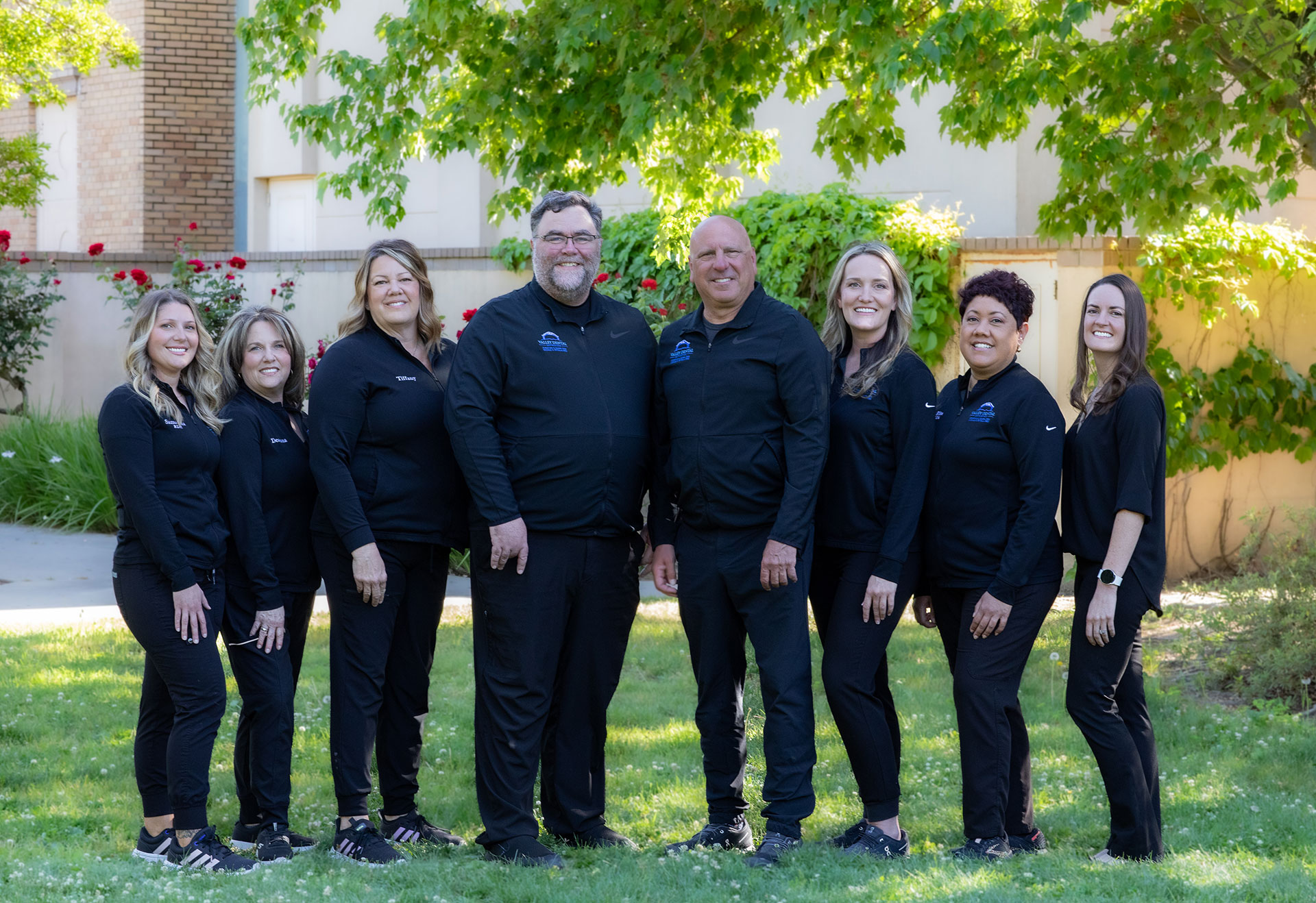 The image shows a group of people, likely a team or group of professionals, posing together for a photograph outdoors. They are dressed in matching black tops with visible logos and white lettering on the front. The individuals appear to be standing in front of a building with a brick facade and a tree behind them.