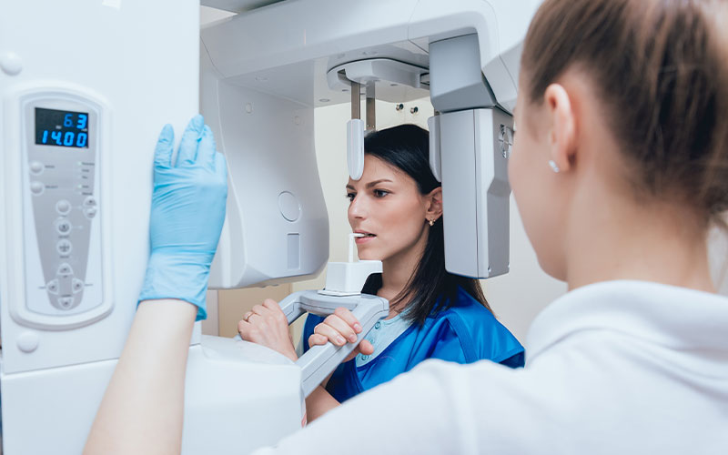 The image shows a woman in a medical gown standing next to a large 3D scanner, which is being operated by another person wearing blue gloves and a white coat. They are both in a room with medical equipment and the environment suggests they are in a medical setting.